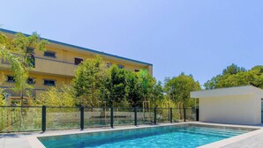 Ein Schwimmbecken mit Glasbarriere, einer kleinen weißen Struktur und einem gelben Gebäude mit blauen Fenstern.