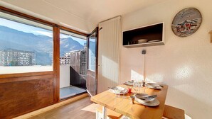 A dining area with a wooden table set for two, a window with a view of buildings and mountains, and a wall-mounted shelf with dishes. - Bright studio, ski-in/ski-out - 4 guests (Les Menuires)