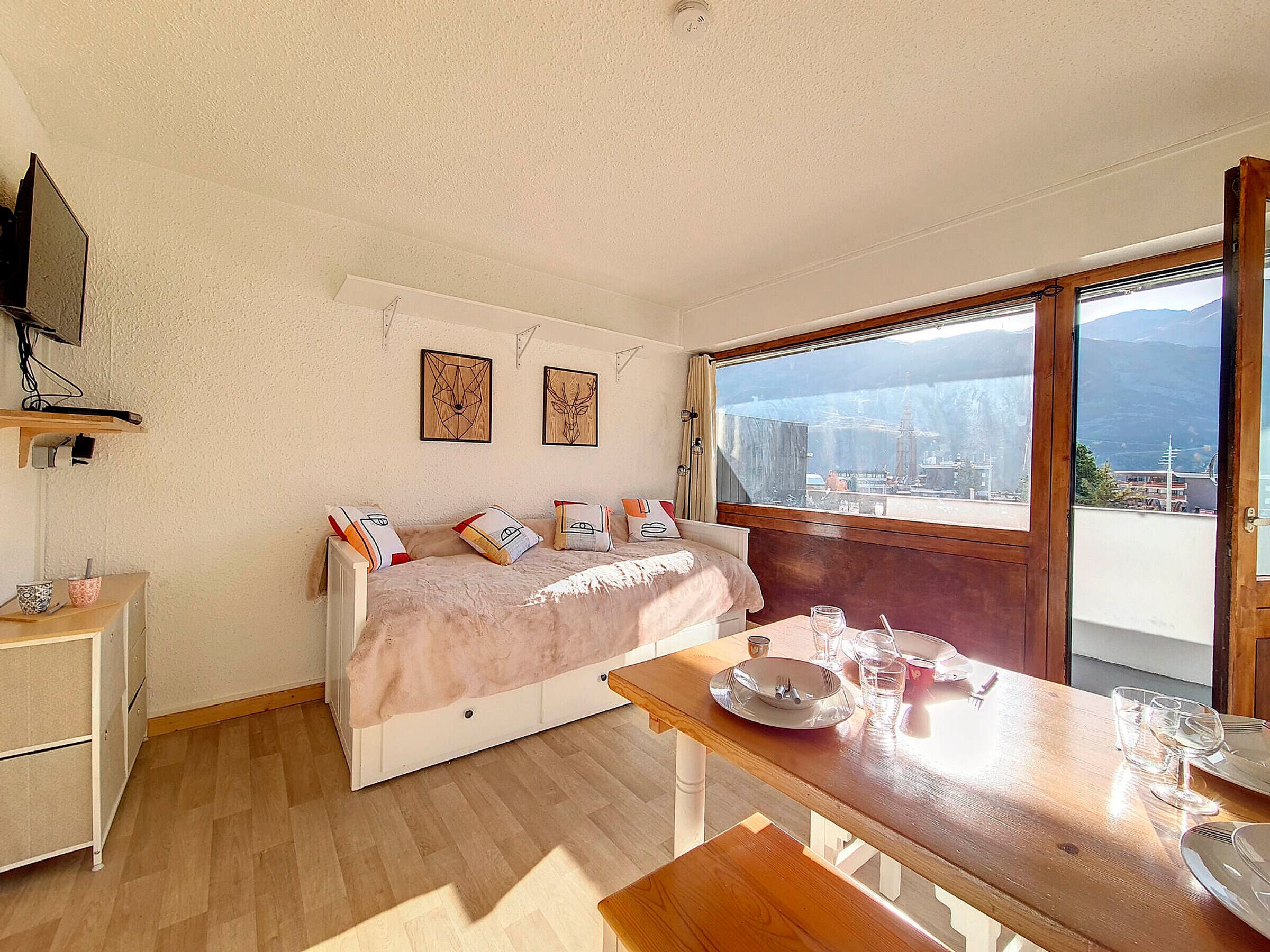 A bedroom with a bed, a table with plates and glasses, a TV on the wall, and a window with a view of buildings and mountains.