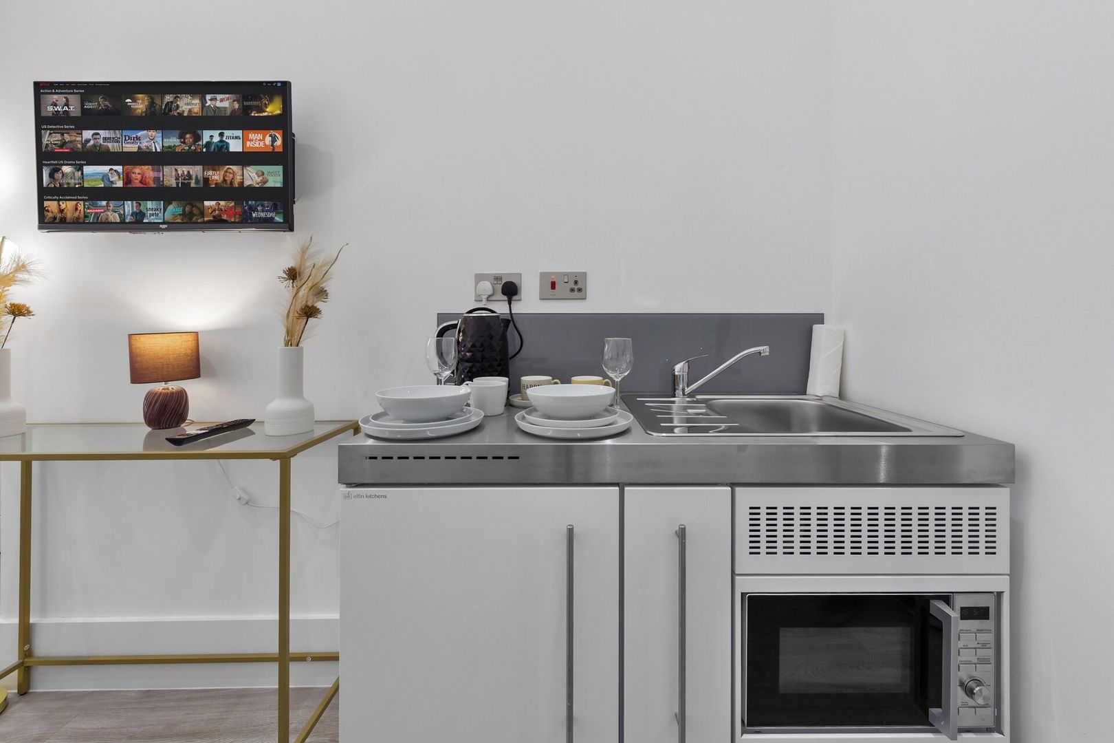 A modern kitchen with a built-in microwave, a stainless steel sink, and a flat-screen TV mounted on the wall displaying various shows.