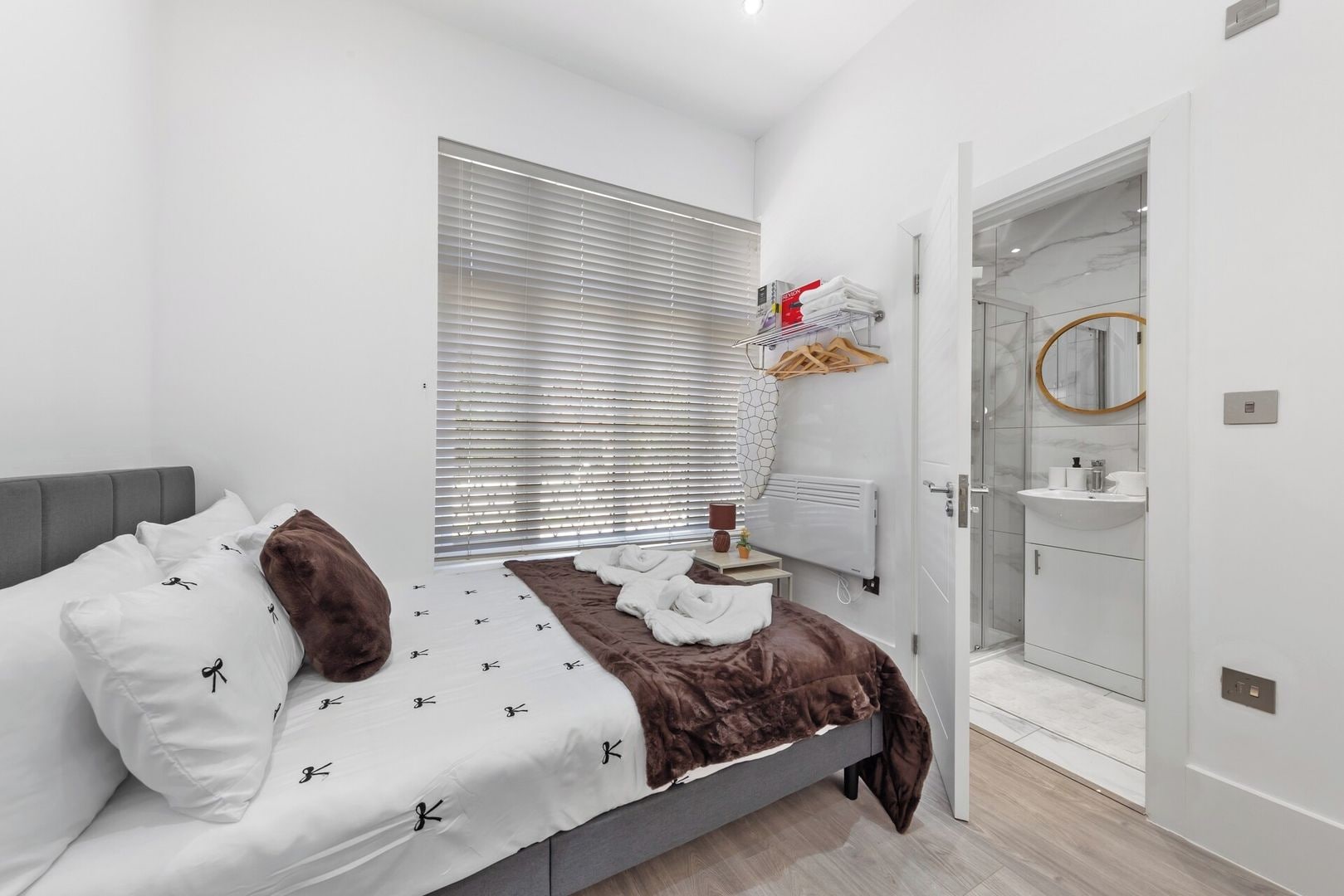 A bedroom with a bed, a bedside table, a window with blinds, a bathroom with a sink and mirror, and a door leading to another room.