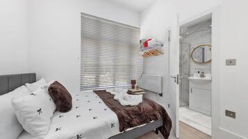 A bedroom with a bed, a bedside table, a window with blinds, a bathroom with a sink and mirror, and a door leading to another room.