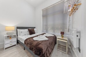 A modern bedroom with a bed, bedside tables, a lamp, a radiator, and a window with blinds.