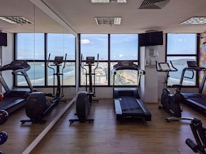 A gym with cardio equipment and a large window overlooking a beach and cityscape. - SHB Magia de uma Vista Panorâmica (Santos)