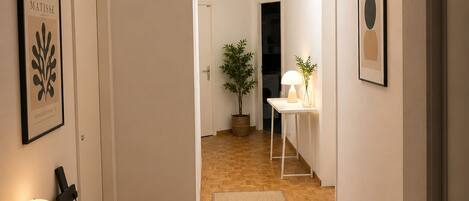 A hallway with a wooden floor, a rug, a desk with a lamp, and framed artwork on the walls.