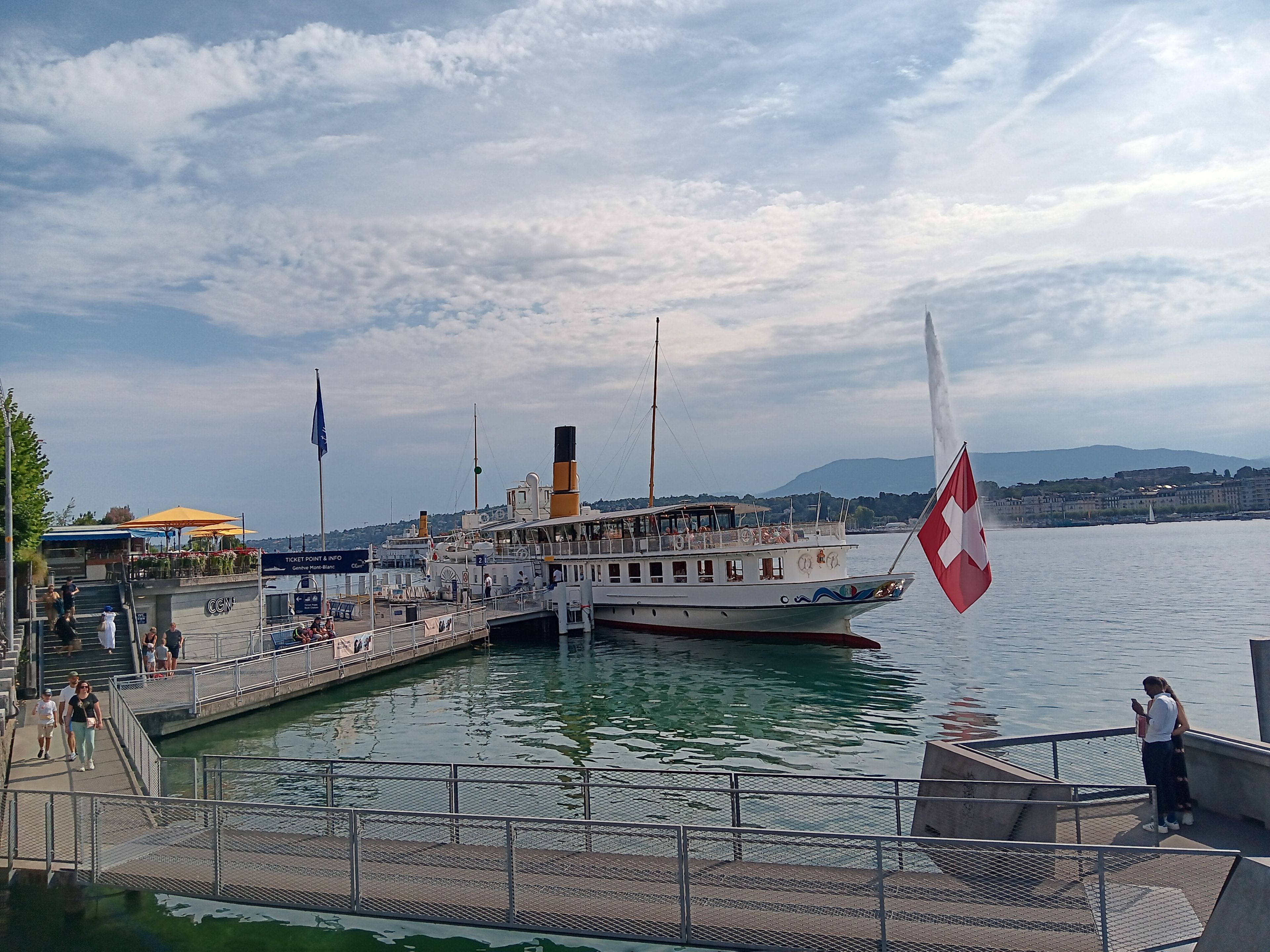 Ein an einem Steg vertäutes Boot mit einer Flagge, eine Prominade mit Personen und eine Stadtlandschaft im Hintergrund.