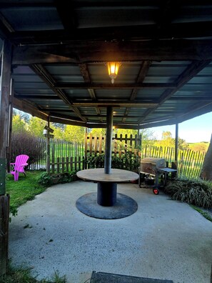 Ein überdachter Außenbereich mit Tisch und Grill, umgeben von Holzzaun und Grünpflanzen.