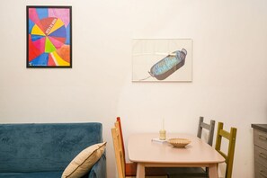 Ein Zimmer mit einem blauen Sofa, einem kleinen Tisch mit einer Kerze und zwei Stühlen. An der Wand hängen zwei gerahmte Kunstwerke.