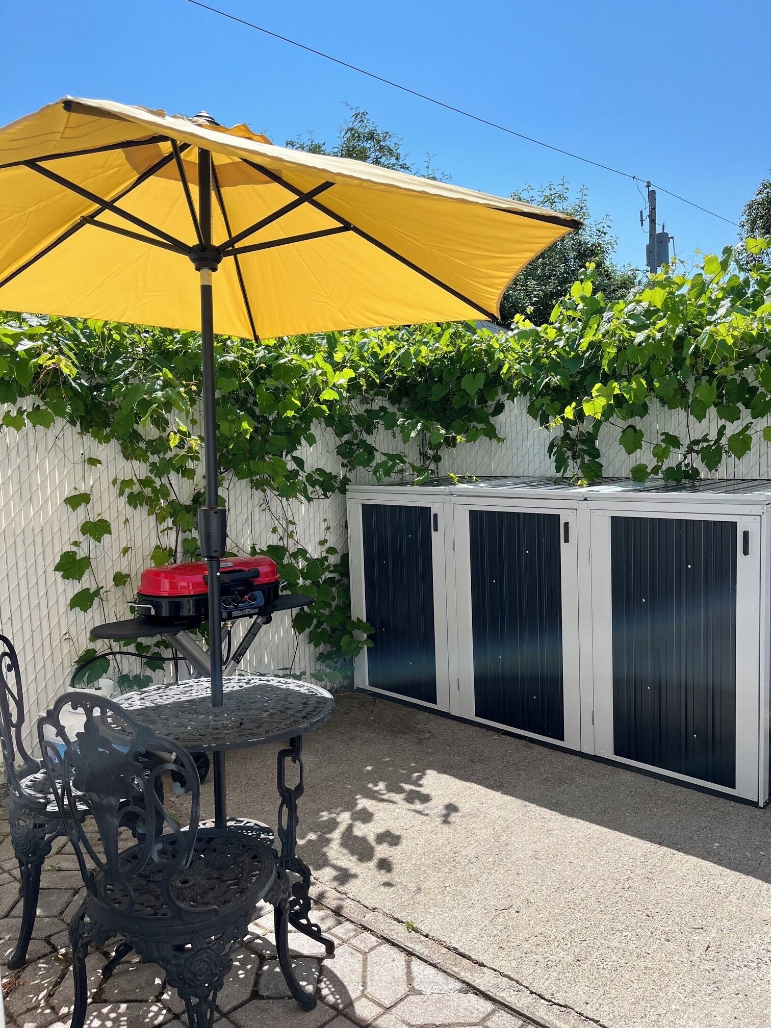 A patio area with a table and chairs, a yellow umbrella, and a white fence with green vines.