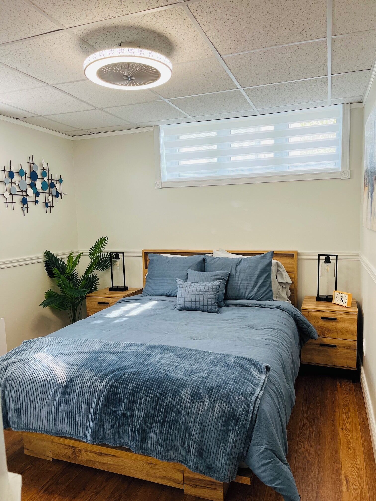 A bedroom with a wooden bed, a nightstand with a clock, a wall-mounted light fixture, and a window with blinds.