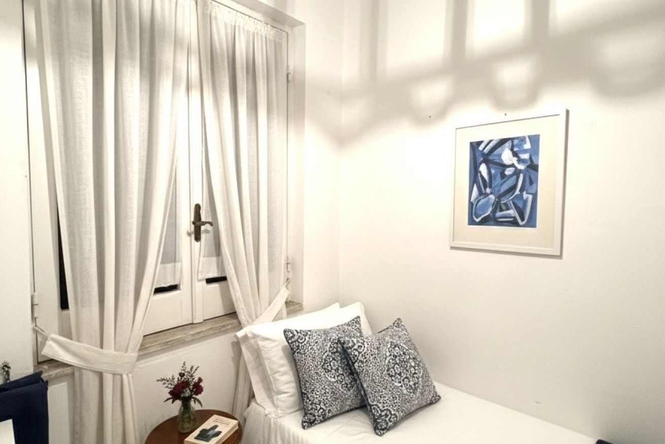 A bedroom with a bed, white curtains, a framed abstract painting, and a vase with flowers.