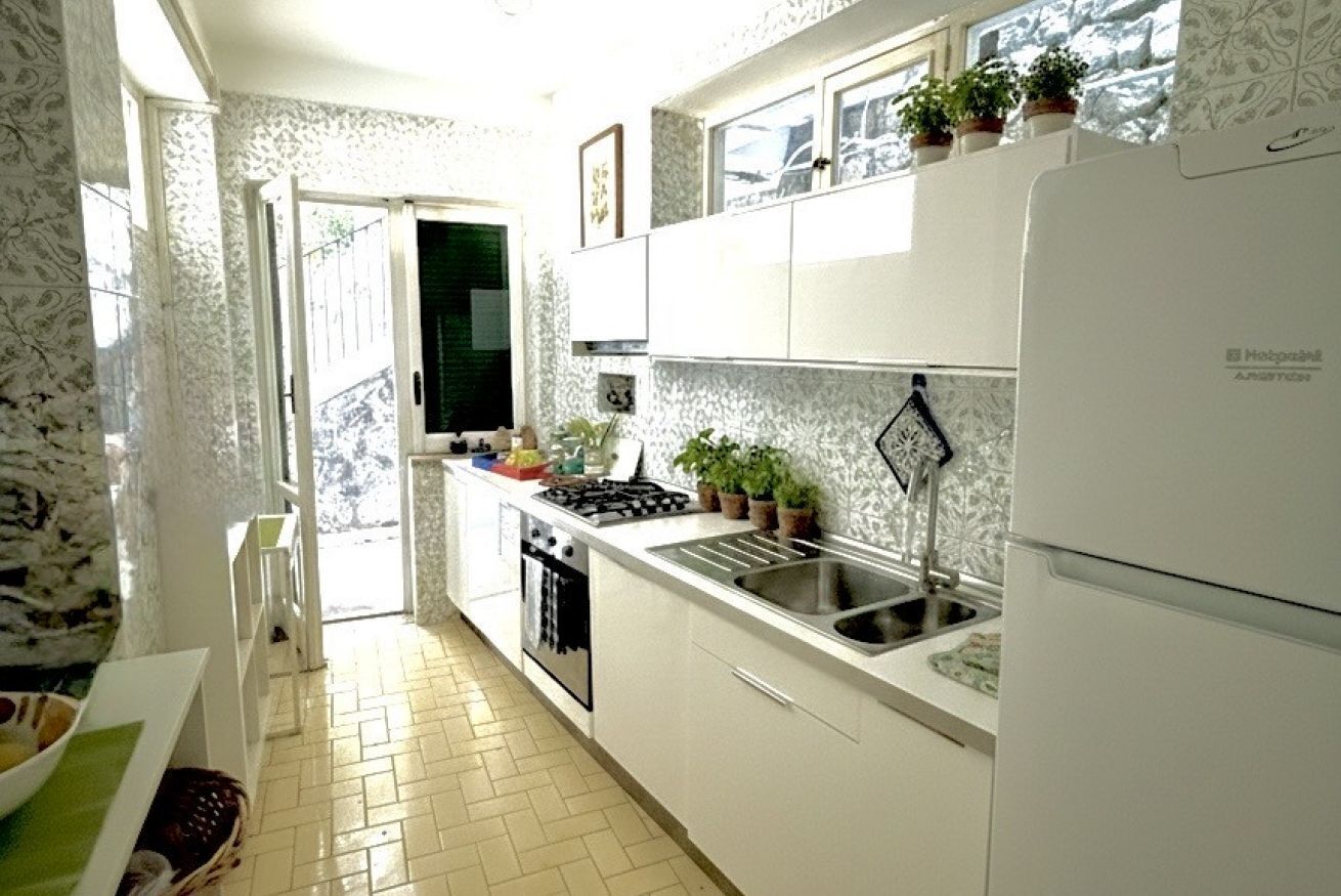 Une cuisine compacte avec des meubles blancs, une cuisinière à gaz, un évier et un réfrigérateur. Des plantes en pot sont posées sur le plan de travail et une fenêtre se trouve au-dessus de l’évier.