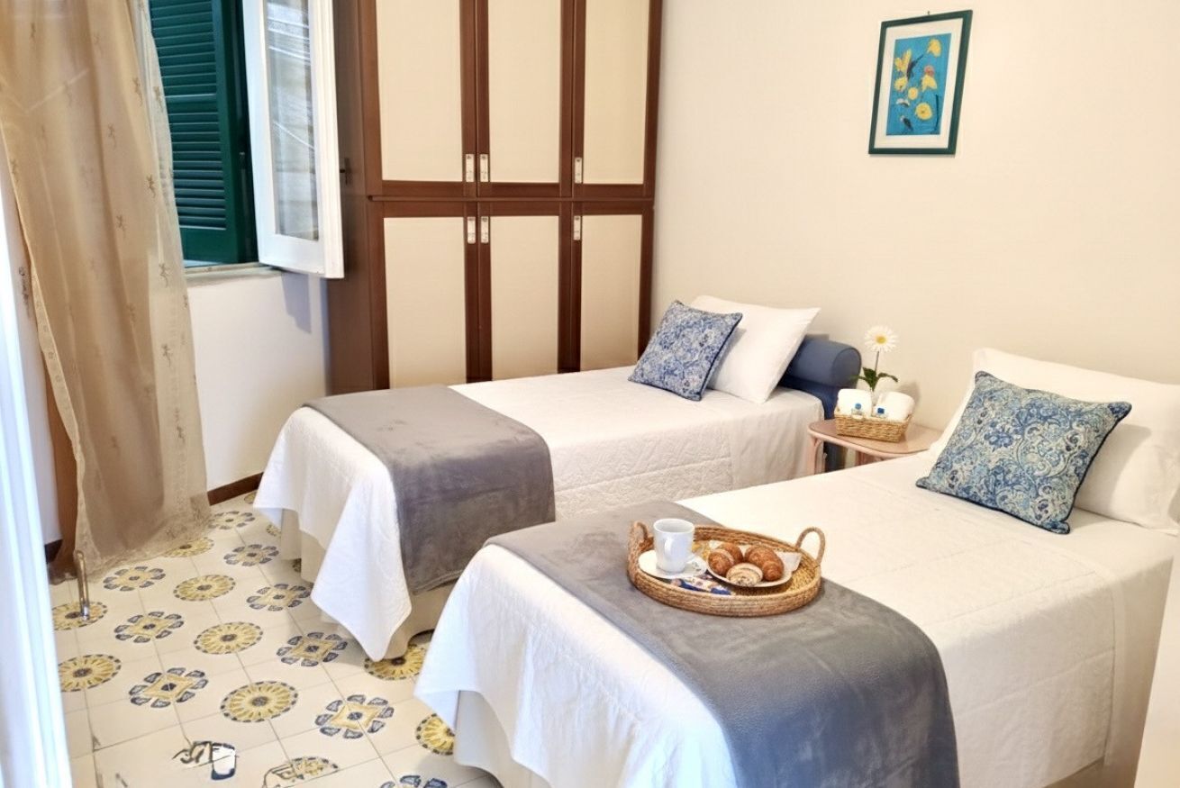 A hotel room with two beds, a small table with a basket of pastries, a window with shutters, and a framed picture on the wall.