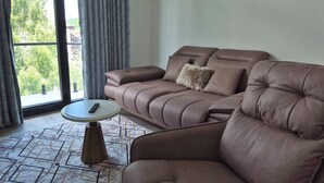 Ein modernes Wohnzimmer mit einem braunen Sofa, einem kleinen runden Tisch und Blick auf Grünflächen außerhalb.