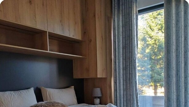 Ein Schlafzimmer mit Bett, Kissen, einer Lampe und Blick auf Bäume durch ein Fenster.