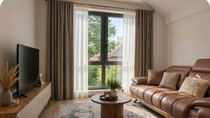 Ein Wohnzimmer mit einer Ledersofa, einem Fernseher auf einem Holzschrank, einem runden Couchtisch und einem großen Fenster mit Vorhängen.