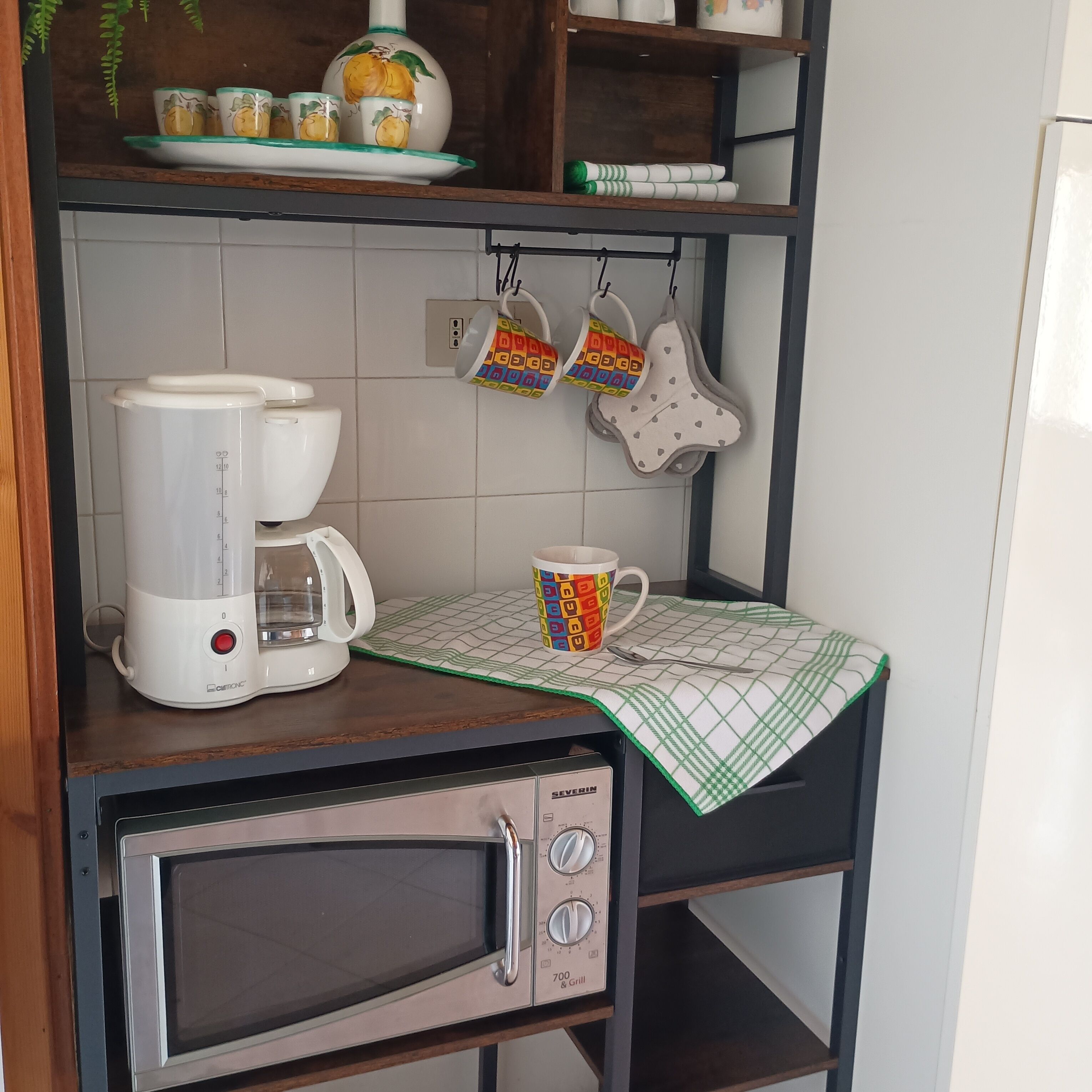 Una cocina con cafetera, microondas y una repisa con tazas y tetera.