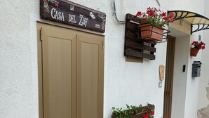 Eine Gebäudefassade mit einem hölzernen Schild über einer geschlossenen Tür, zwei Blumenkästen an der Wand und einem kleinen Fenster mit einem Holzklappladen.