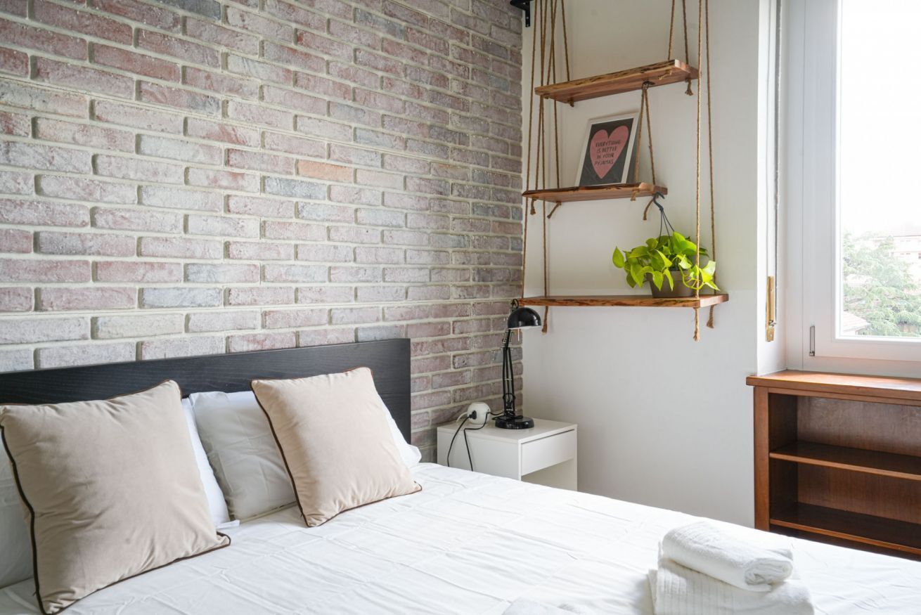 Ein Schlafzimmer mit Backsteinwand, einem Bett mit weißer Bettwäsche, einem Nachttisch mit Lampe und einem Fenster mit Blick.
