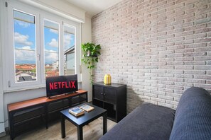 A living room with a brick wall, a TV on a wooden stand, a black cabinet, a grey sofa, and a window with a view of buildings. - Torino Centro | Casa Matilda (Torino)