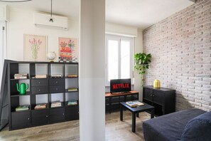 Ein Wohnzimmer mit Bücherregal, einer schwarzen Couch, einem Couchtisch, einem Fernseher, der 'NETFLIX' zeigt, und einer Ziegelwand.
