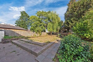 A backyard with a wooden fence, a grassy area, and a variety of trees including a tall weeping willow. - Harlesden Haven Studio (London)