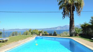 Una piscina rectangular con agua azul cristalina, rodeada por una terraza de piedra, una palmera y vistas al mar y las montañas al fondo.