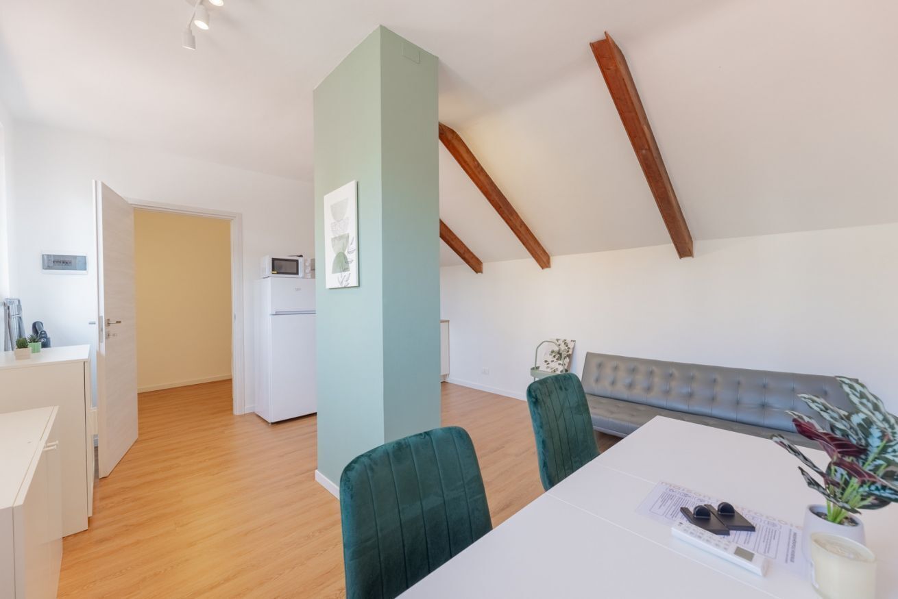 Een moderne keuken met witte kasten, een witte koelkast en een magnetron. Er is een eethoek met een witte tafel en groene fauteuils. Een grijze bank staat tegen de muur en er staat een plant op de tafel.