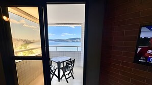 Ein Zimmer mit Blick auf ein Gewässer und Hügel, mit Fliesenboden, einem kleinen Tisch mit Stühlen und einem an der Wand befestigten Fernseher.