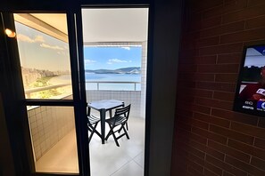 A room with a view of a body of water and hills, a tiled floor, a small table with chairs, and a television on the wall. - SHB Horizonte Azul Uma Janela para o Mar (Santos)