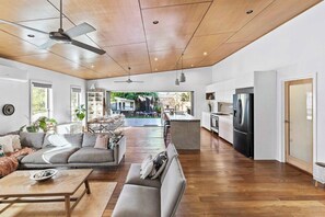A spacious living area with a sofa, a coffee table, and a dining table. There is a kitchen area with a refrigerator and a dining table. The room has a wooden ceiling and large windows.