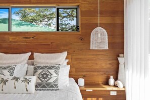 A bedroom with a wooden headboard, a bed with white bedding and patterned pillows, a hanging light fixture, and a view of trees through a window.
