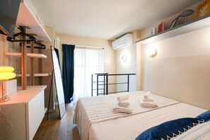A hotel room with a bed, a desk with a lamp, a window with curtains, an air conditioning unit, and a shelf with items. - Cit Turin | Casetta Sogni (Torino)