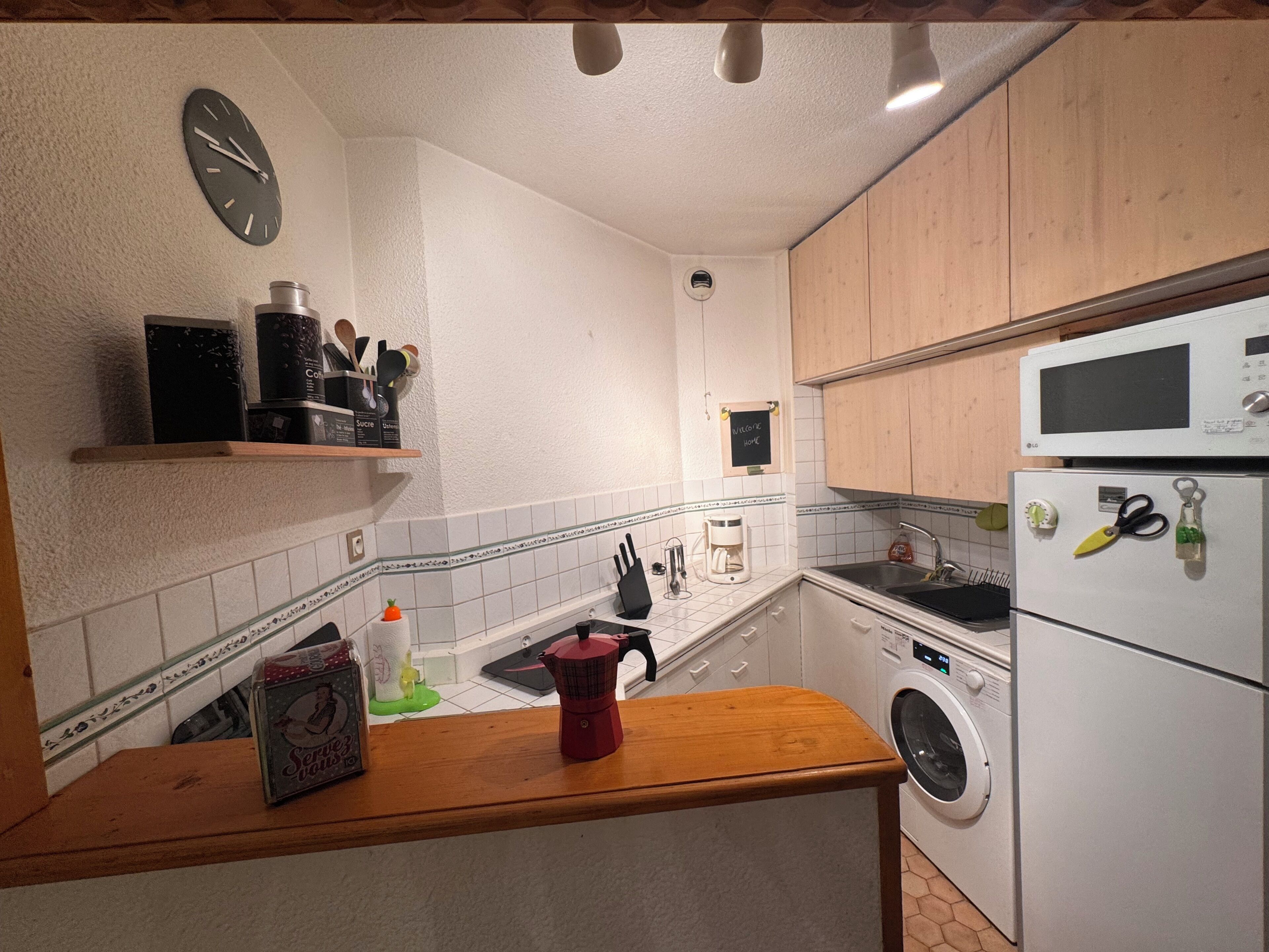 Una cocina con un reloj, una cafetera, una lavadora y un microondas.