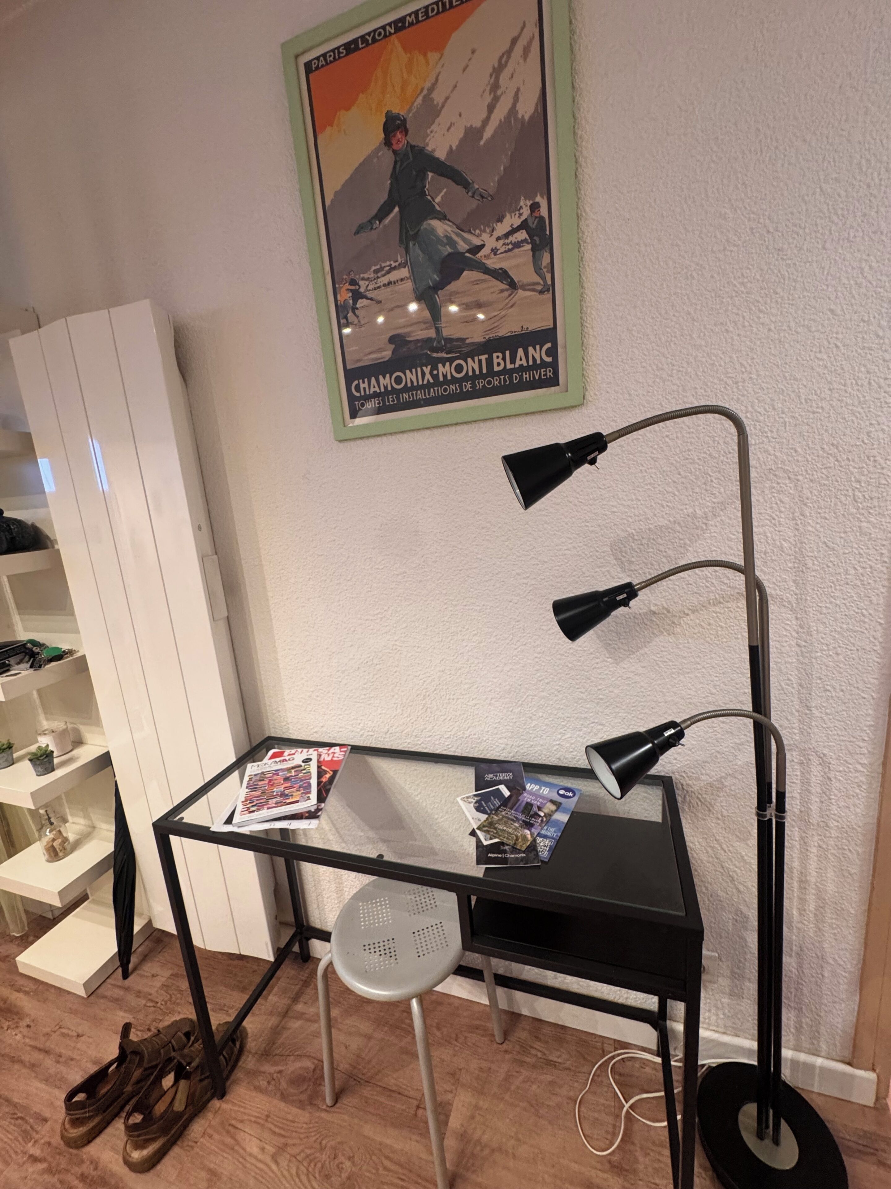 A room with a desk, a chair, a standing lamp, a poster on the wall, and a shoe rack.