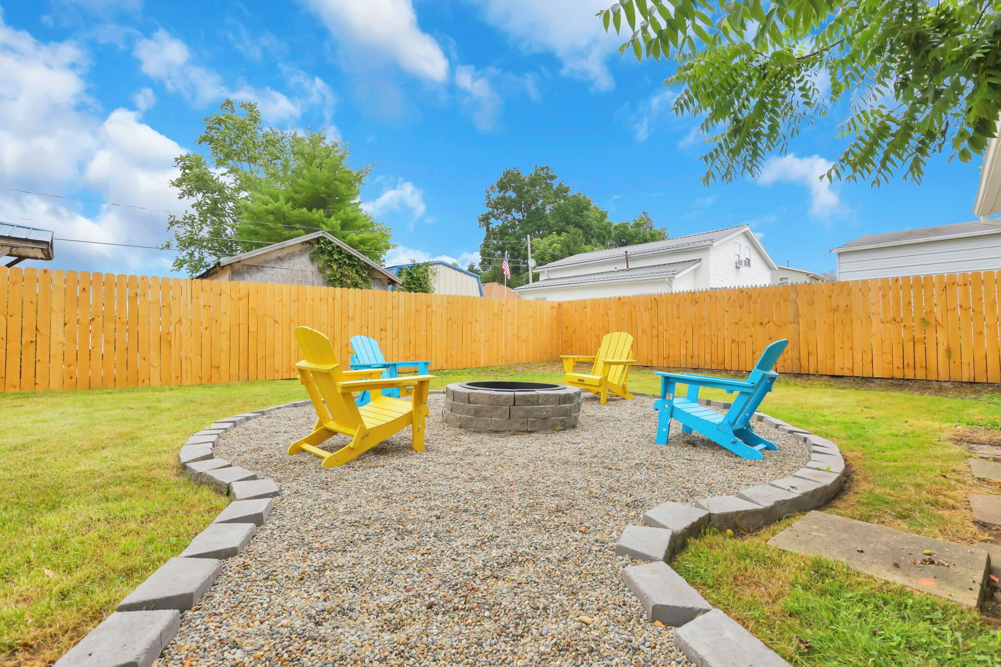 A backyard with a wooden fence, a circular fire pit, and two Adirondack chairs.