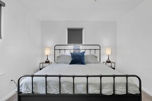Ein Bett mit Metallgestell, zwei Nachttische mit Lampen, ein Fenster über dem Bett und eine schlichte weiße Wand.