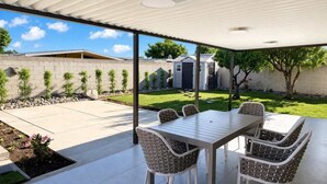 Une terrasse couverte avec une table et des chaises, une pelouse, un mur en briques et un petit abri de jardin.