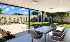 Eine überdachte Terrasse mit Tisch und Stühlen, ein Rasen, eine Ziegelmauer und ein kleines Gartenhaus.