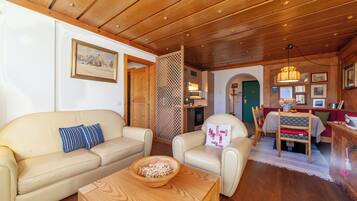 Una sala de estar acogedora con techo de madera, un sofá, una silla, una mesa con un cuenco y un comedor con una mesa y sillas.