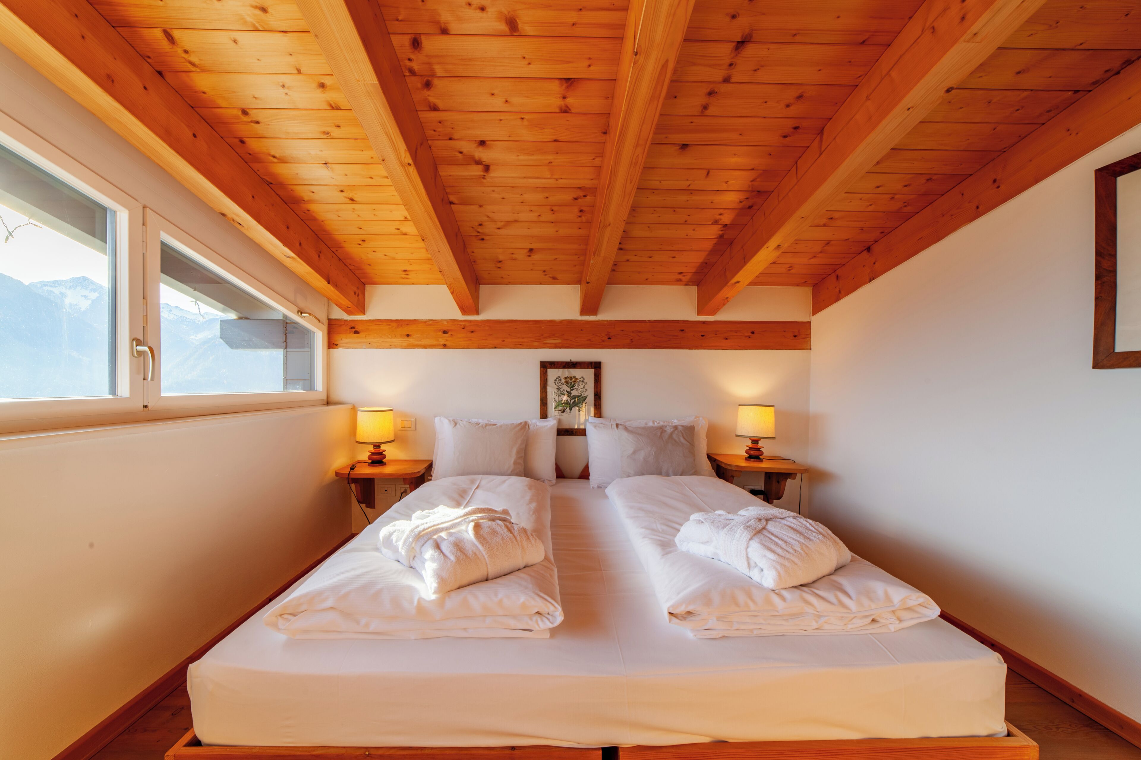 A double bed with white linens and towels, wooden beams on the ceiling, bedside lamps, and a window with a view.