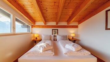 A double bed with white linens and towels, wooden beams on the ceiling, bedside lamps, and a window with a view.