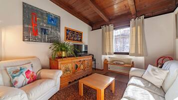 A living room with a wooden ceiling, a sofa, a coffee table, a TV, a radiator, and a window with curtains.