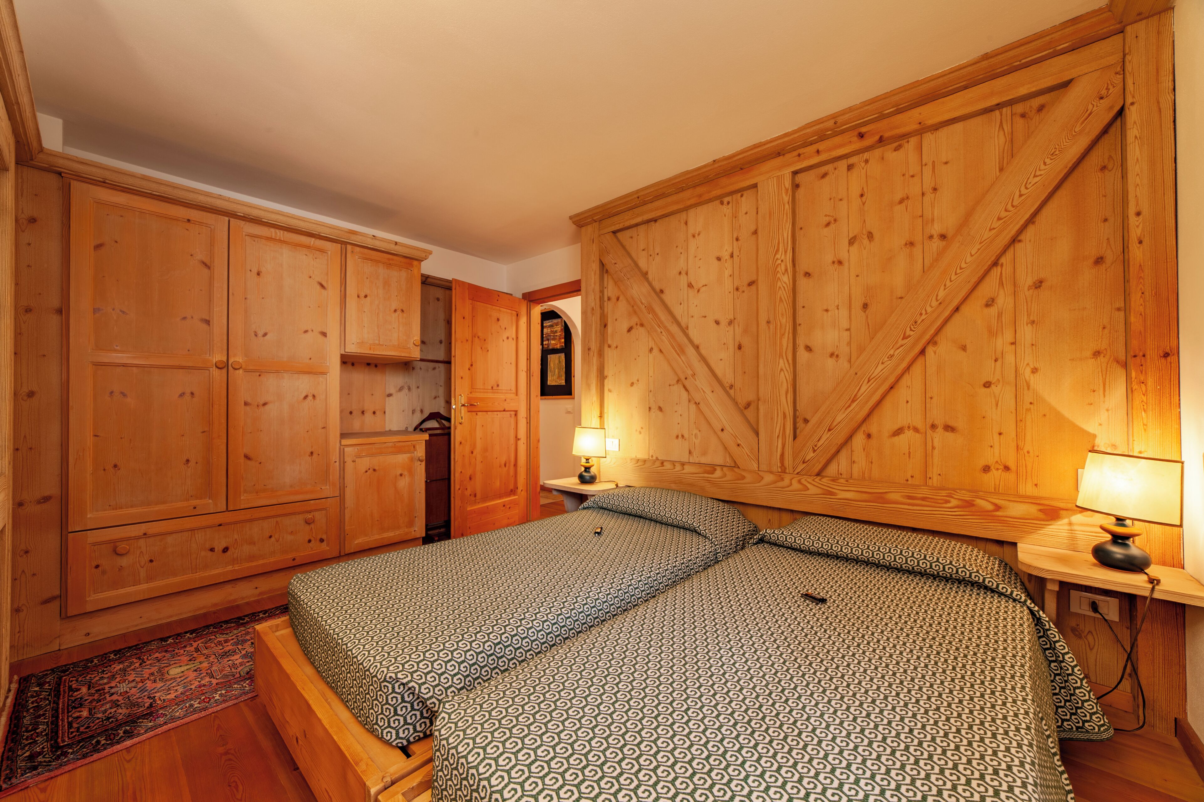 A bedroom with a large bed, wooden wardrobe, and a small table with a lamp.