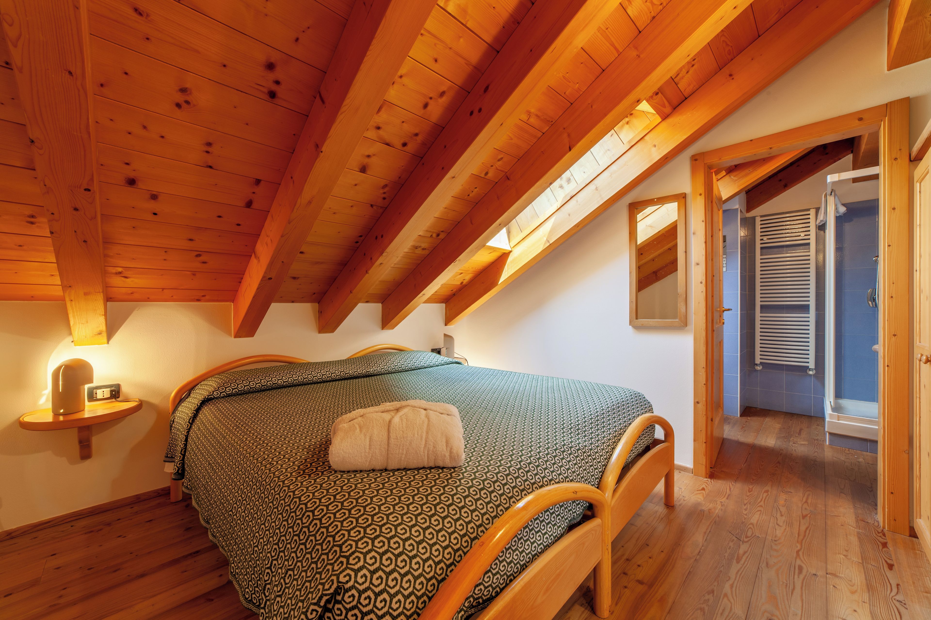Un dormitorio con techo de madera, una cama con ropa de cama estampada, una mesita de noche con una lámpara y una puerta que da a un baño.