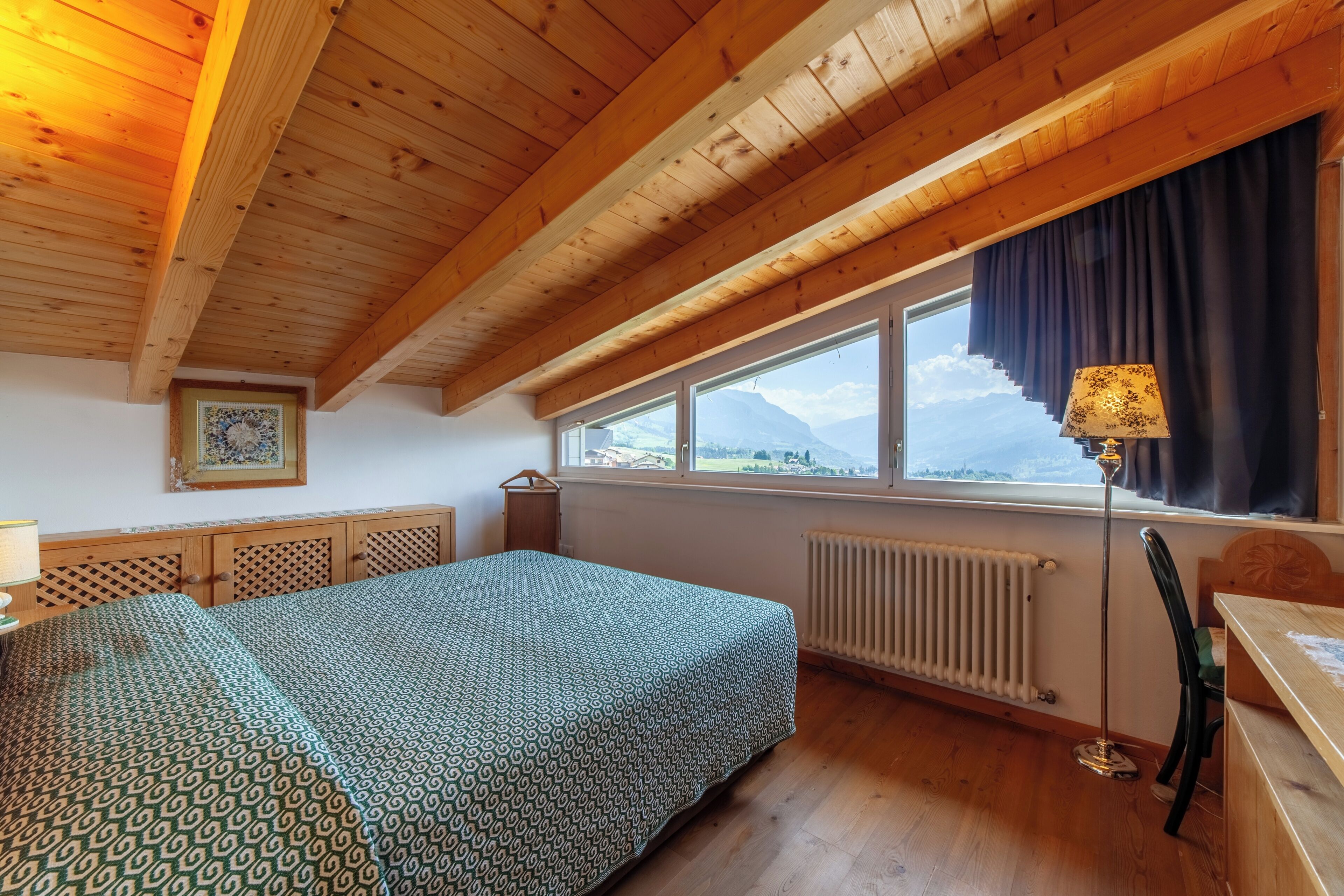 Ein Schlafzimmer mit Holzboden, einem Bett mit gemustertem Überwurf, einem Fenster mit Bergblick, einem Heizkörper und einer Lampe.