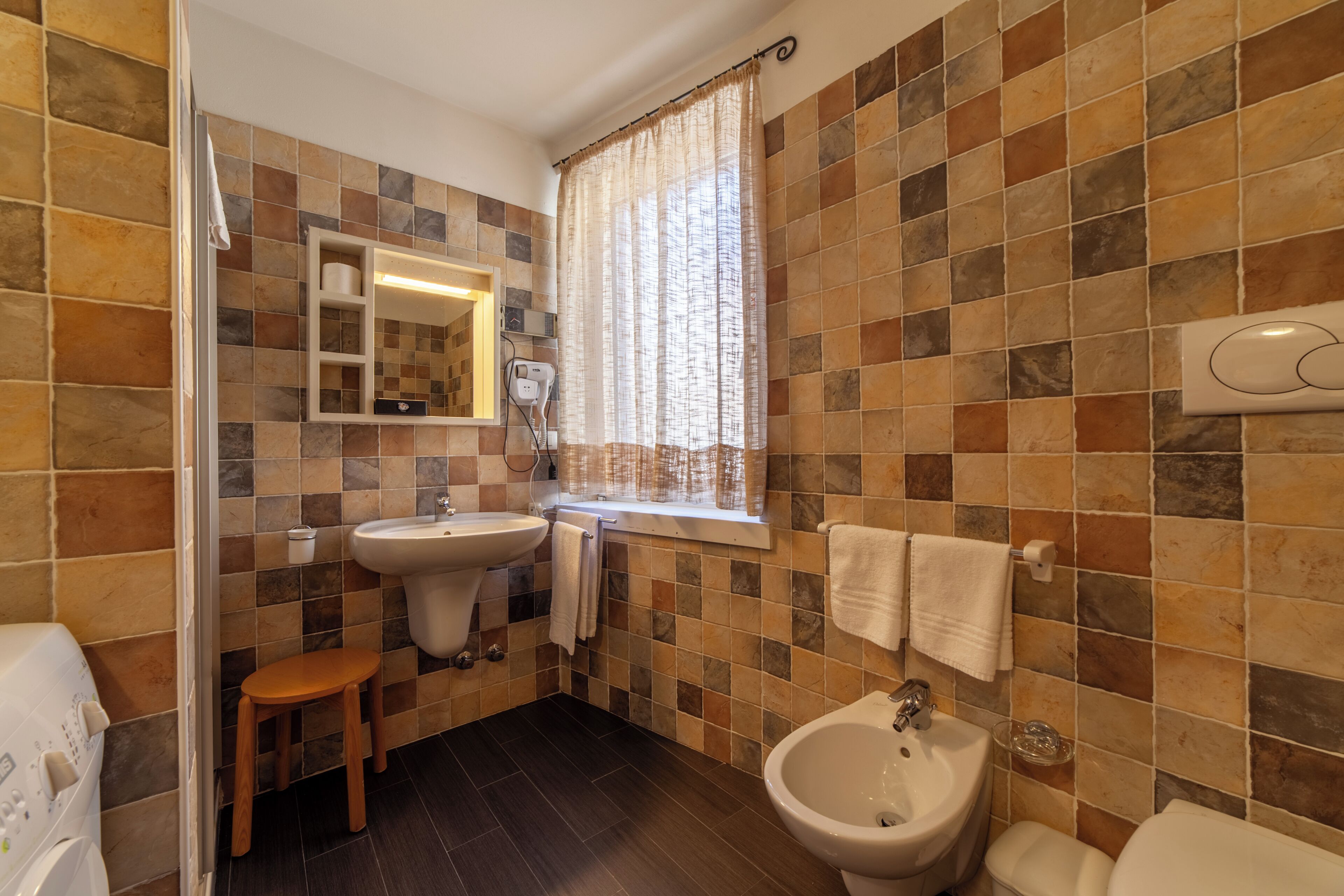 Une salle de bain comprenant un lavabo, un bidet et des toilettes, avec des murs carrelés et une fenêtre avec des rideaux.