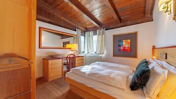 A bedroom with a wooden ceiling, a bed with white bedding, a desk with a chair, a mirror, and a window with curtains.