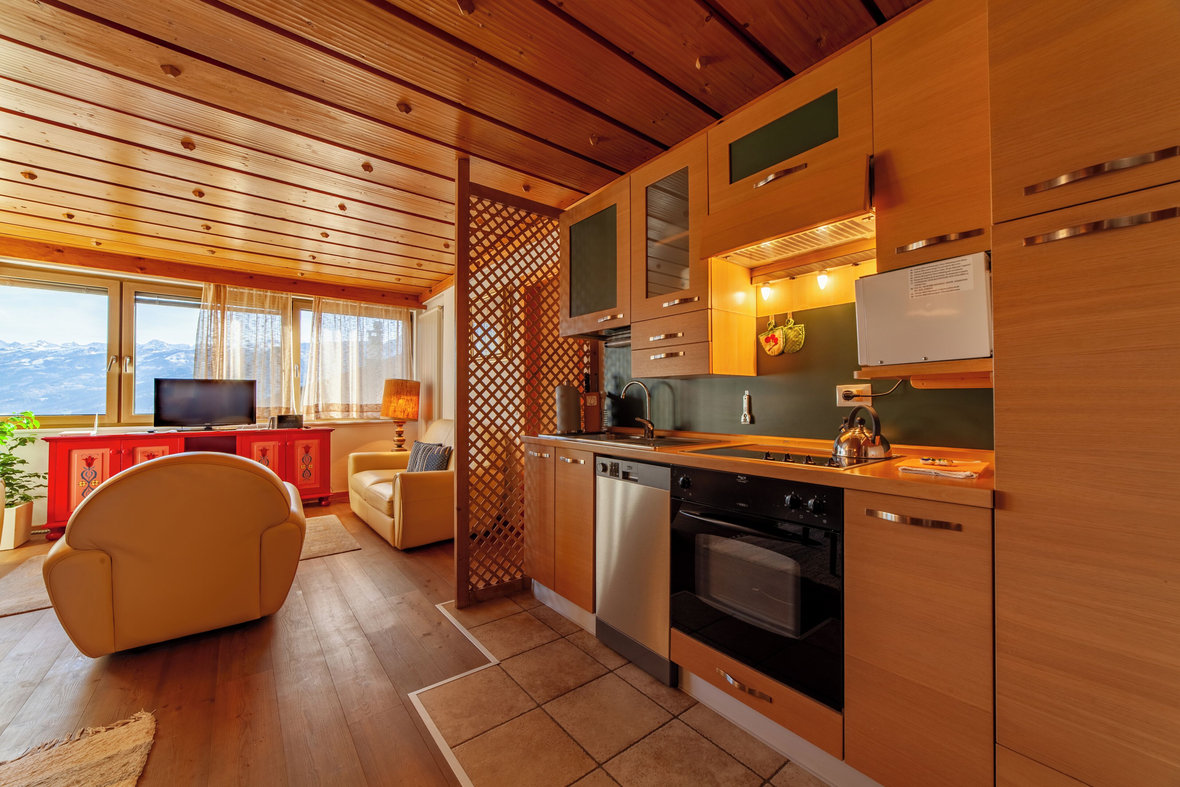Een moderne keuken met houten kastjes, een vaatwasser en een fornuis. In de woonkamer staat een televisie, met uitzicht op bergen door het raam.
