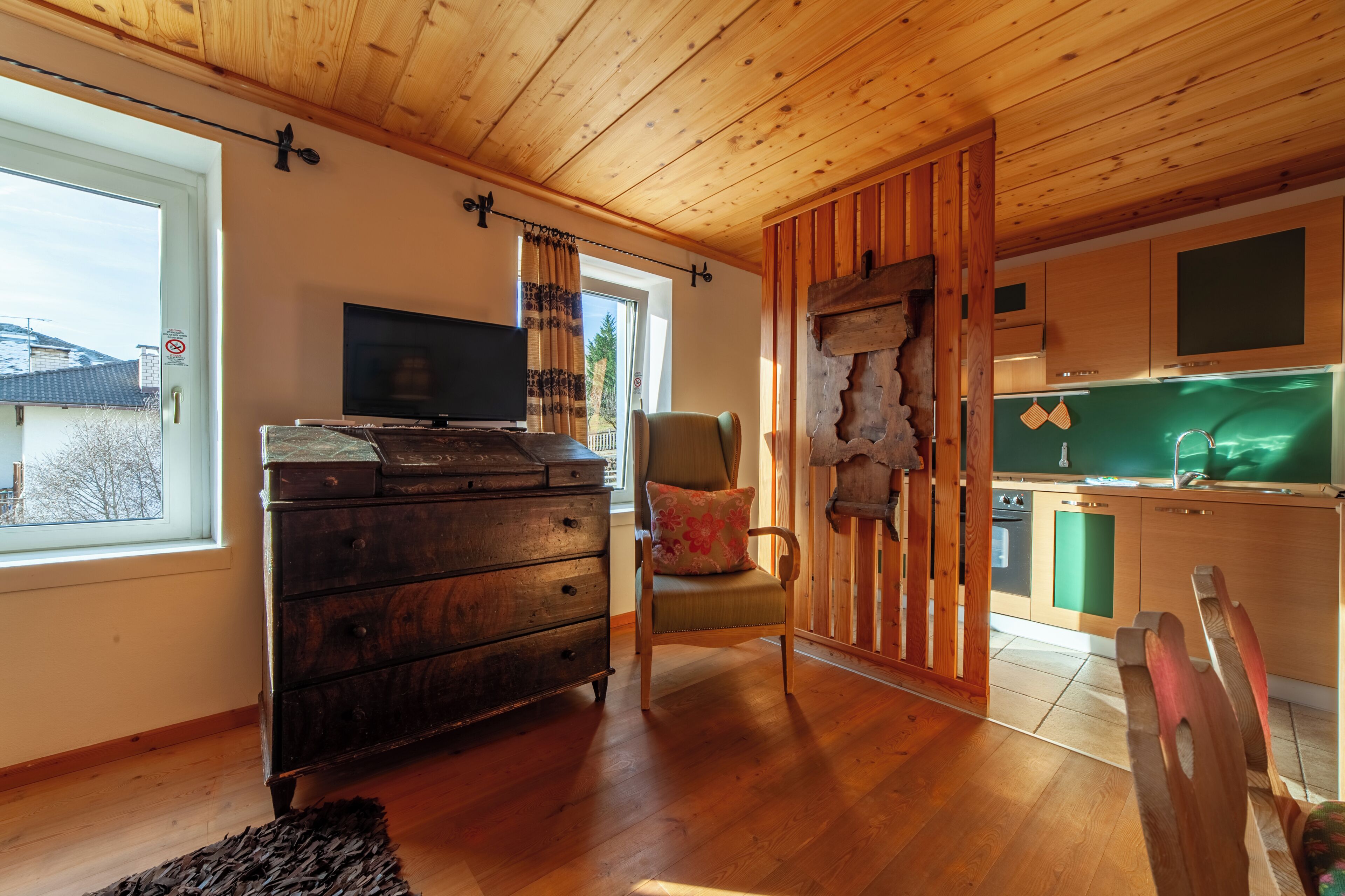 Una habitación acogedora con techo de madera, televisor, cómoda de madera, una silla y una zona de cocina con un mueble verde y un lavamanos.